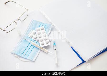 Medizinischer Hintergrund: Pillen, Brille, Stift, Maske, Thermometer, Impfstoff, Spritze und Papier mit Patientendaten. Medizinisches Thema. Konzept: Impfung gegen Coronavirus. Stockfoto