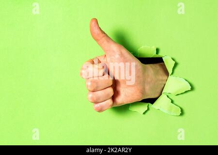 Hand mit Daumen nach oben durch Loch in grüner Papierwand Stockfoto