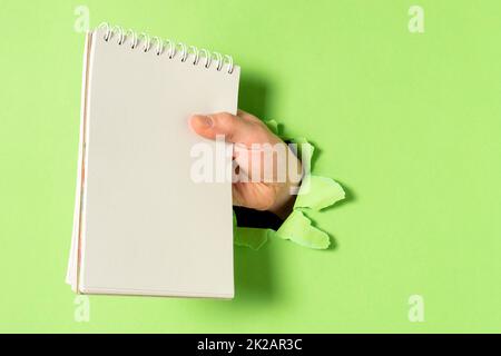 Hand mit Spiralnotizbuch durch Loch in grüner Papierwand Stockfoto