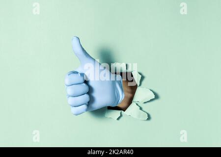 Hand in Medizinhandschuh mit Daumen-hoch-Geste durch Loch in Papierwand Stockfoto