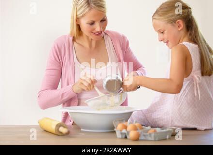 Misch alles in mit Mama. Ein kleines Mädchen, das auf einer Küchentheke sitzt und ihrer Mutter beim Backen hilft. Stockfoto