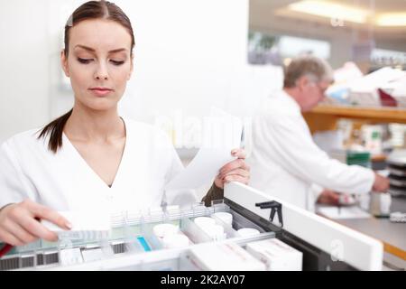 Apotheker prüft Medikamente mit Kollegen. Porträt einer Apothekerin, die Medikamente überprüft, mit einer Kollegin, die im Hintergrund arbeitet. Stockfoto