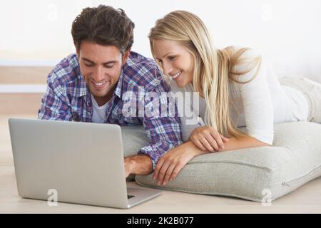 Brauchen Sie eine neue Couch, die nur einen Klick entfernt ist. Ein glückliches junges Paar, das zu Hause auf dem Boden liegt und zusammen einen Laptop benutzt. Stockfoto