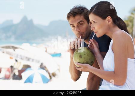 Ich genieße etwas Neues mit der Person, die ich liebe. Das junge Paar genießt einen Drink von einer Kokosnuss, während es einen Tag am Strand genießt. Stockfoto