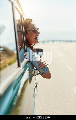 Sich auf offener Straße frei fühlen. Eine kurze Aufnahme einer attraktiven Frau, die beim Roadtrip aus einem Autofenster hängt. Stockfoto