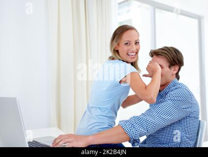 Kein Blick. Eine junge Frau bedeckt die Augen ihres Freundes, während er an einem Laptop arbeitet. Stockfoto