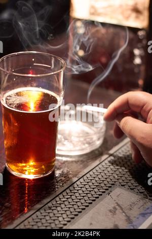 Sich schlechten Gewohnheiten hingeben. Kurzer Schuss einer mans-Hand, die eine Zigarette und ein Glas Bier auf der Theke hält. Stockfoto