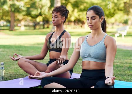 Sich auf Achtsamkeit und den Atem konzentrieren. Eine kurze Aufnahme von zwei attraktiven jungen Frauen, die nebeneinander sitzen und im Park meditieren. Stockfoto