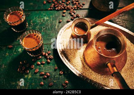 Frisch gebrühter orientalischer Kaffee in den Cizves und Gläser, die in der hellen Küche auf einem schäbigen grünen Tisch liegen Stockfoto