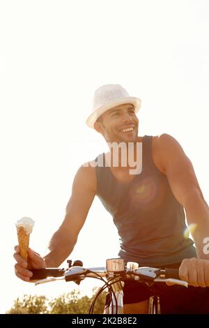 Genießen Sie einen Eiskegel und eine Radtour. Aufnahme eines hübschen jungen Mannes, der ein Eis isst, während er mit dem Fahrrad im Freien fährt. Stockfoto