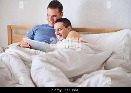 Ein junges schwules Paar, das ein digitales Tablet benutzt, während es sich im Bett entspannt. Stockfoto