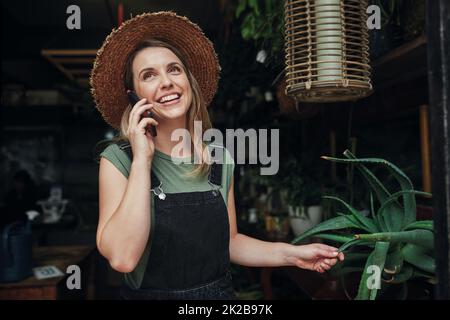 Ich liebe es, positives Feedback von einem Kunden zu erhalten. Ein Bild einer attraktiven jungen Geschäftsinhaberin, die allein am Eingang ihrer Blumengeschichte steht, während sie telefoniert. Stockfoto