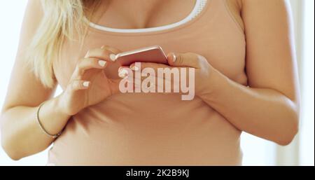 Mit allen in Kontakt bleiben vor dem großen Tag. Ausgeschnittene Aufnahme einer unkenntlich werdenden Schwangeren, die zu Hause ein Handy benutzt. Stockfoto