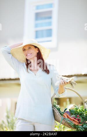 Im Garten zu arbeiten ist, wie ich mich erhole. Eine schöne Frau hält ihre Gartenhandschuhe und einen Korb mit frisch gepflücktem Gemüse, während sie in ihrem Garten steht. Stockfoto