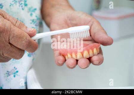 Ältere oder ältere Patienten in Asien verwenden eine Zahnbürste, um Teilprothesen von Ersatzzähnen zu reinigen. Stockfoto