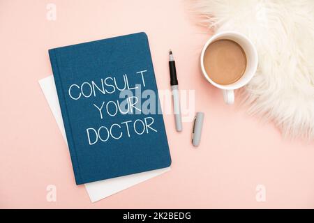 Textzeichen mit „Arzt konsultieren“. Konzeptfoto Fragen Sie Informationen oder Rat von einem medizinischen Fachmann Bürobedarf über Schreibtisch mit Tastatur und Brille und Kaffeetasse für die Arbeit Stockfoto