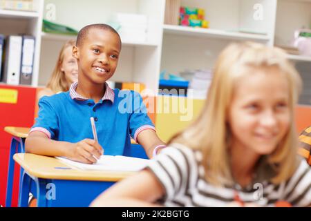 HES macht das Beste aus seiner Ausbildung. Junger afroamerikanischer Student, der unter Klassenkameraden an seinem Schreibtisch lächelt - Copyspace. Stockfoto