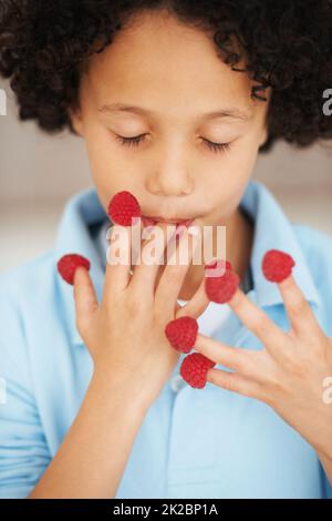 Er kann gar nicht genug von diesen Himbeeren bekommen. Ein netter kleiner Junge, der Himbeeren mit geschlossenen Augen von den Fingern isst. Stockfoto