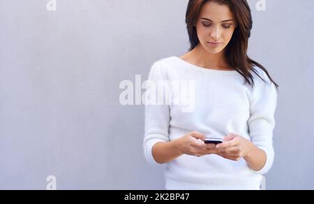 Geduldig auf seine Antwort warten. Aufnahme einer attraktiven jungen Frau, die vor grauem Hintergrund steht und ihr Mobiltelefon benutzt. Stockfoto