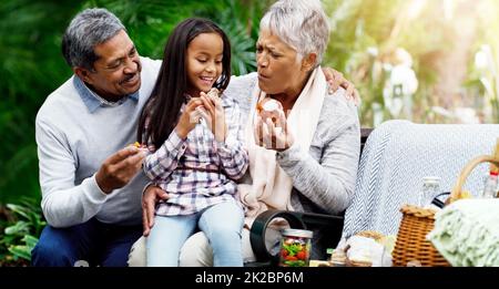 Wir haben immer gesunde Leckereien für sie. Aufnahme eines fröhlichen kleinen Mädchens, das mit ihren Großeltern im Park auf einer Bank sitzt. Stockfoto