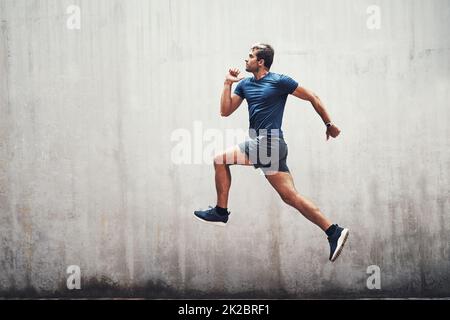 Halten Sie die Energie hoch. Aufnahme eines sportlichen jungen Mannes, der draußen gegen eine graue Wand läuft. Stockfoto