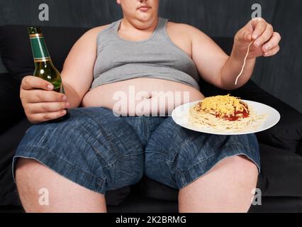 Wer braucht eine Gabel. Eine ausgeschnittene Aufnahme eines übergewichtigen jungen Mannes, der auf einem Sofa sitzt und Spaghetti mit den Händen isst. Stockfoto