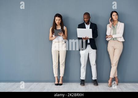 Sie sind damit beschäftigt, ihre Beziehungen erfolgreich zu gestalten. Aufnahme einer Gruppe von Geschäftsleuten, die digitale Geräte nutzen, während sie vor grauem Hintergrund in der Schlange stehen. Stockfoto