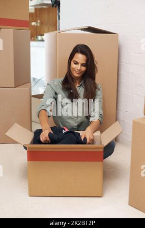 Und so beginnt das Auspacken. Eine schöne junge Frau, die in ihrem neuen Haus auf dem Boden sitzt und Kartons auspacket. Stockfoto