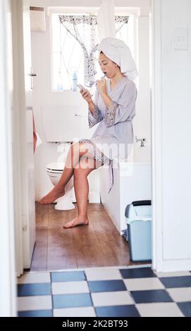 Senden eines Kurztextes während der Auffrischung. Aufnahme einer jungen Frau, die ihr Handy benutzte, während sie sich im Badezimmer die Zähne putzte. Stockfoto