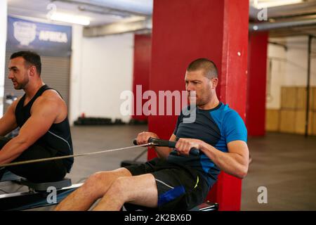 Rudern, um zerrissen zu werden. Eine kurze Aufnahme von zwei Männern, die im Fitnessstudio am Rudergerät trainieren. Stockfoto