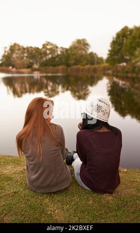 Gute Zeit mit meinem Partner in der Kriminalität. Rückansicht von zwei Mädchen im Teenageralter, die neben einem See sitzen und sich unterhalten. Stockfoto