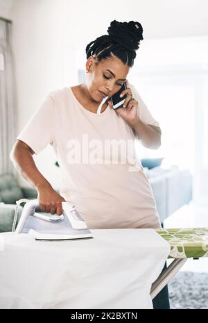 Multitasking ist eine Fähigkeit. Aufnahme einer jungen Frau, die zu Hause am Telefon Kleidung bügelt. Stockfoto