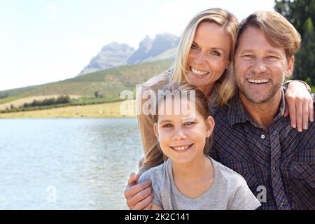 Erinnerungen, die ein Leben lang andauern. Porträt einer glücklichen dreiköpfigen Familie, die an einem See sitzt. Stockfoto