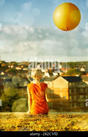Ihre Kindheit wiedererleben. Rückansicht einer jungen Frau, die mit einem Ballon und einer Stadt im Hintergrund sitzt. Stockfoto