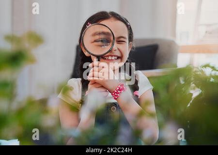 Alles sieht größer aus. Ein Porträt eines entzückenden kleinen Mädchens, das lächelt, während es zu Hause durch eine Lupe schaut. Stockfoto