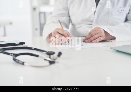 Einige Patientenunterlagen werden sortiert. Ausgeschnittene Aufnahme einer nicht erkennbaren Ärztin, die während ihrer Arbeit in ihrem Büro auf ein Dokument schreibt. Stockfoto