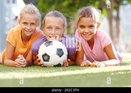 Unsere Schule bietet Mädchen Fußball. Drei lächelnde Mädchen, die auf sonnigem Gras liegen und mit einem Fußball-Copyspace posieren. Stockfoto