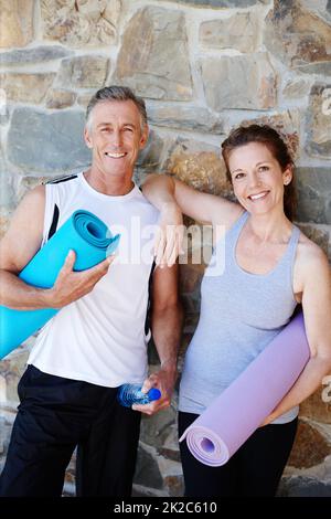 Sie lieben ihre Yoga-Kurse. Ein reifes Paar, das seine Yogaausrüstung mit einem breiten Lächeln im Freien hält. Stockfoto