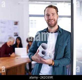 Ein aufsteigender Stern im Architekturspiel. Porträt eines jungen Architekten mit Blaupausen und im Hintergrund arbeitenden Kollegen. Stockfoto