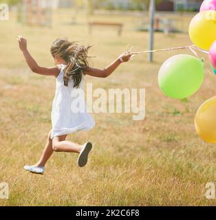 Glücklich hüpfend. Rückansicht eines kleinen Mädchens, das mit einer Reihe von Luftballons draußen durch ein Feld läuft. Stockfoto