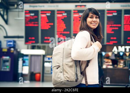 Ich freue mich schon auf die Reise. Zugeschnittenes Porträt einer attraktiven jungen Frau, die an einem Ankunfts- und Abflugsbrett im Flughafen steht. Stockfoto