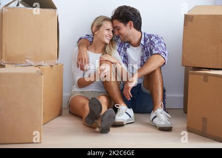 Glücklich in ihrem neuen Liebesnest. Ein glückliches junges Paar, das in ihrem neuen Zuhause auf dem Boden sitzt und kuschelt, während es von Kisten umgeben ist. Stockfoto