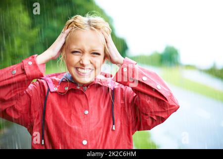 Nichts ist besser als ein wenig frisches Regenwasser. Wunderschöne junge blonde Frau trägt einen roten Regenmantel im Regen draußen auf einer Landstraße. Stockfoto