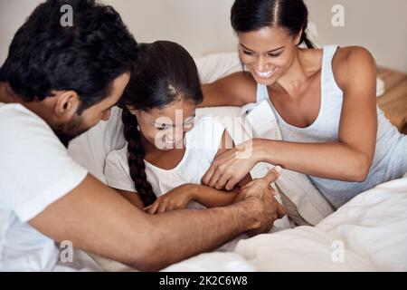 Zeit für dummen Spaß. Aufnahme von Eltern, die ihre kleine Tochter kitzeln, während sie zu Hause im Bett liegen. Stockfoto