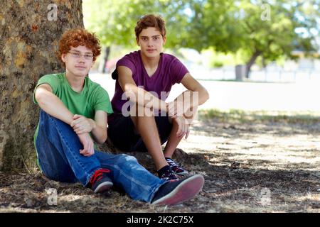 An einem heißen Tag etwas Schatten fangen. Aufnahme von zwei Jungen im Teenageralter, die sich im Schatten eines Baumes im Park entspannen. Stockfoto