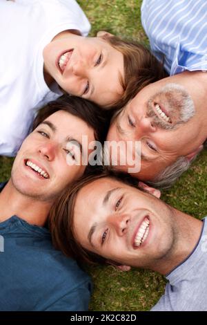 Gemeinsam durch die Zeitalter lachen. Vater und Söhne liegen auf dem Gras, ihre Köpfe nebeneinander. Stockfoto