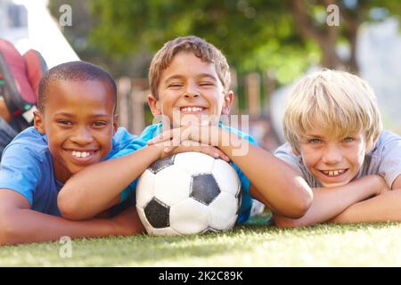 Ein gewinnendes Team aus besten Freunden. Drei glückliche Jungen, die mit ihrem Soccerball - Copyspace draußen in der Sonne liegen. Stockfoto