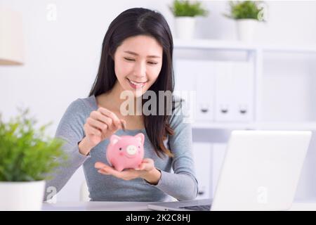 Junge Frau, die im Heimbüro Münzen in die Sparschwein legt Stockfoto