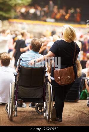 Mein Handicap hindert mich nicht daran, das Leben zu genießen. Rückansicht einer Frau, die neben ihrem Mann steht, der bei einer Live-Musikveranstaltung im Rollstuhl sitzt. Stockfoto