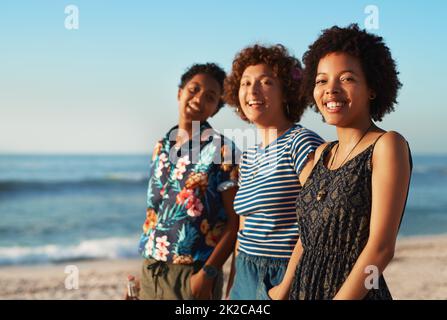 Wir lassen uns gegenseitig lächeln. Porträt von drei attraktiven jungen Frauen, die tagsüber zusammen stehen und am Strand posieren. Stockfoto